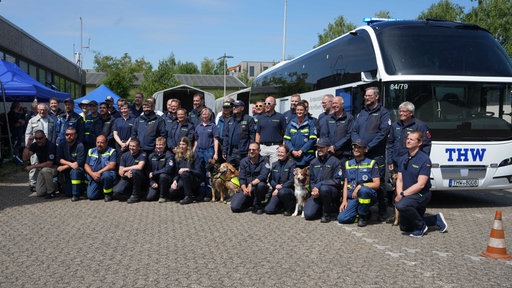 Eine Gruppe von Menschen in blauer THW-Kleidung, in der Mitte ein Hund, dahinter ein THW-Bus.