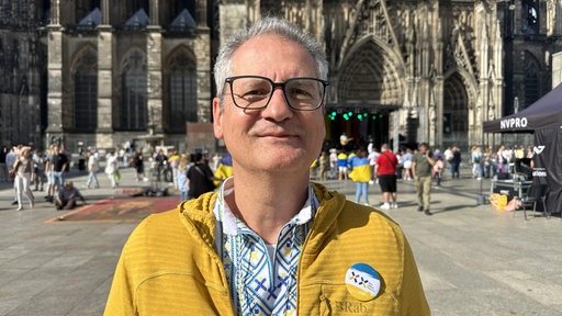 Mann mit grauen Haaren und Brille trägt eine gelbe Jacke und einen blau-weiß-gelben Pin