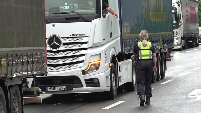Ein weißer LKW wird von einer Polizistin auf der Autobahn gestoppt, die zum Fenster am Fahrhaus läuft. 
