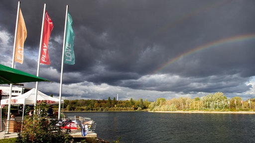 An einem Seeufer steht ein Pavillion und am Himmel sind dunkle Wolken und ein Regenbogen zu sehen