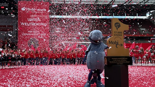Konfetti fliegt durch die Luft im Rhein-Energie-Stadion. Im Vordergrund ein Preis mit der Aufschrift 17 000 Kinder und ein Maskottchen daneben. Im Hintergrund viele Kinder.