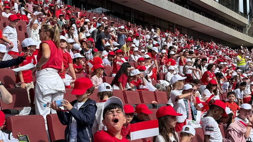 Schüler, Schülerinnen und Lehrpersonal singen im Kölner Rhein-Energie Stadion, schwingen rot-weiße Fahnen.