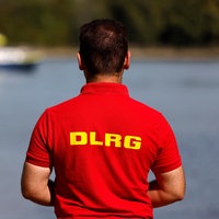 Ein Mitarbeiter der DLRG steht am Rheinufer, im Hintergrund ein Boot der Wasserschutzpolizei