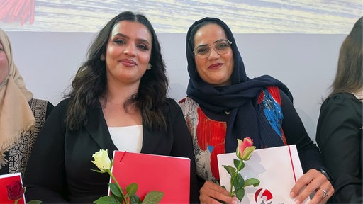 Zwei Frauen halten jeweils ihr Abschlusszeugnis und eine Rose in der Hand und lächeln in die Kamera.