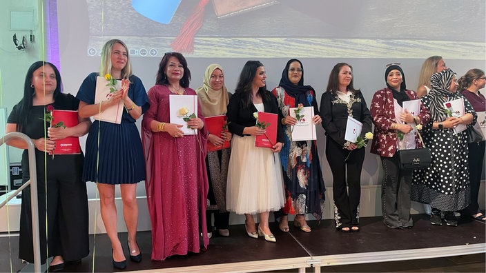 Auf einer Bühne stehen Frauen mit ihren Abschlusszeugnissen und jeweils einer Rose in der Hand.