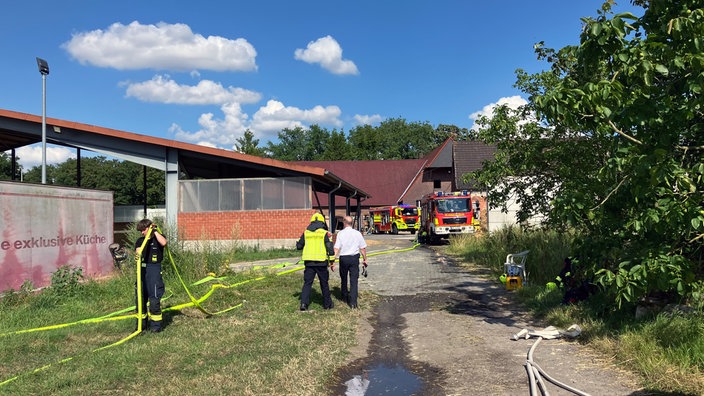 Zwei Feuerwehrwagen stehen auf einem Hof, ein Feuerwehrmann trägt einen Wasserschlauch.