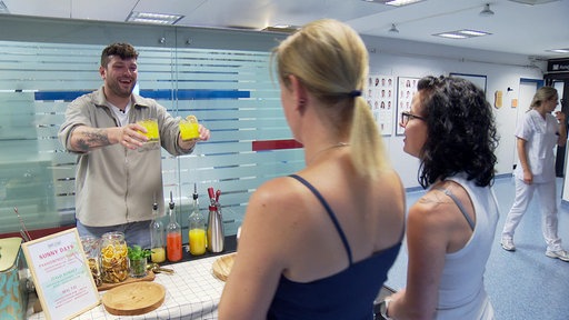 Eine aufgebaute Bar im Krankenhausflur, ein Barkeeper reicht zwei Frauen zwei Drinks.