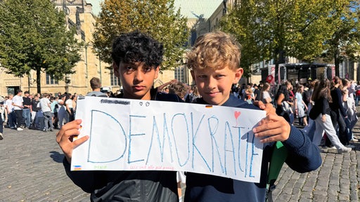 Zwei Jungen halten ein Schild hoch auf dem Demokratie steht.