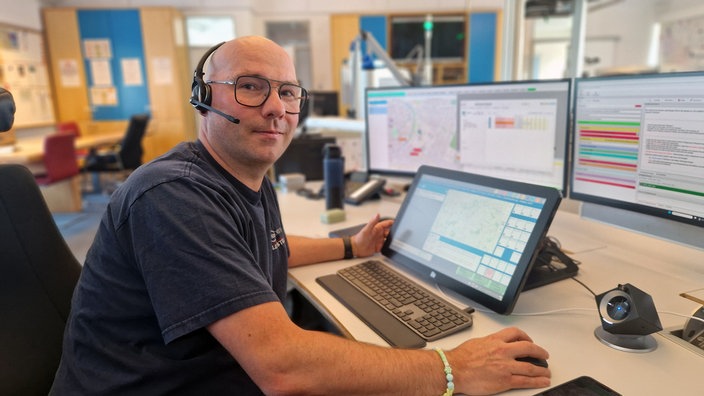 Ein Mann sitzt in einem Büroraum vor Computerbildschirmen und hat ein Headset auf.