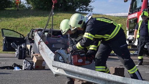 Zwei Feuerwehrmänner zerschneiden eine Leitplanke. Im Hintergrund das Unfall-Auto.