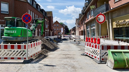 Ein Bagger steht hinter einem Bauzaun auf einer aufgerissenen Straße.