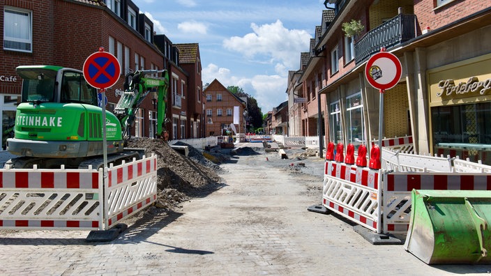 Ein Bagger steht hinter einem Bauzaun auf einer aufgerissenen Straße.