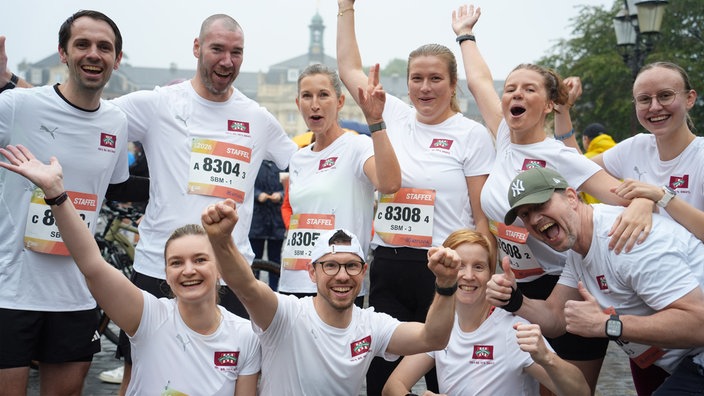 Zehn Läuferinnen und Läufer jubeln in Gruppenpose vor dem Schloss in Münster. Sie tragen weiße T-Shirts und Startnummern.