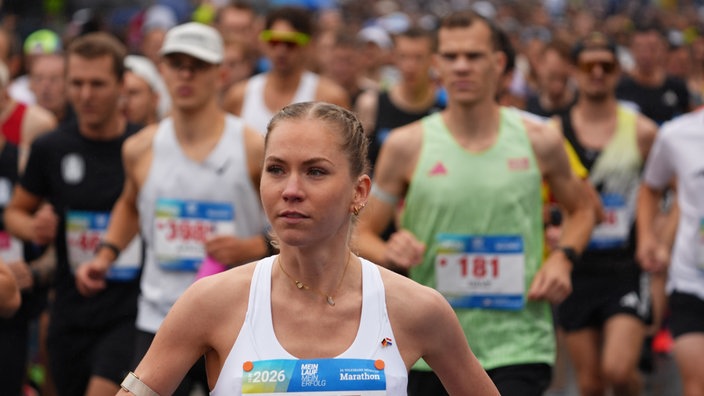 Eine Marathon-Läuferin mit weißem Tanktop läuft an der Kamera vorbei. Hinter ihr sind viele andere Läufer zu sehen.