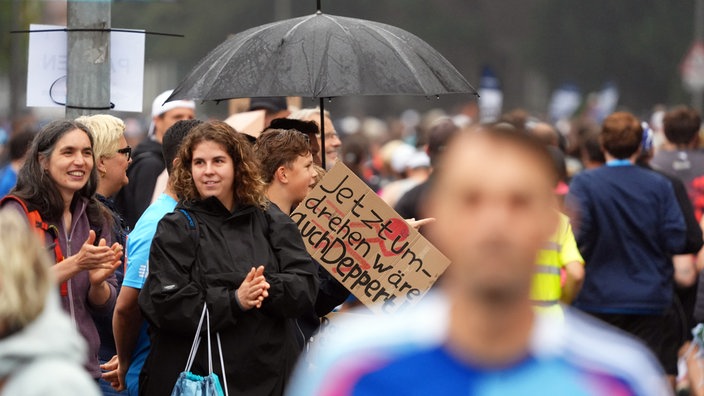 Am Rande der Marathon-Strecke stehen Zuschauerinnen und Zuschauer. Zwei Frauen klatschen, daneben ein braunes Schild mit der schwarzen Aufschrift "Jetzt umdrehen wäre deppert"