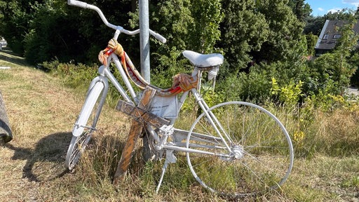 Ein weißes Fahrrad an einem Pfosten mit einem Holzkreuz davor.