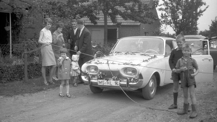 Alte Schwarz/Weiß-Aufnahme in Nienberge aus dem Jahr 1960 oder 1970. Es stehen Hochzeitsgäste um ein Auto herum. Die vorne stehen halten ein gespanntes Seil vor dem Auto.
