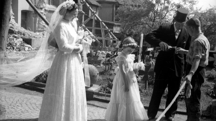 Alte Schwarz/Weiß-Aufnahme von einer Hochzeit in Nienberge aus dem Jahr 1957.  Eine Braut mit Blumenmädchen und Bräutigam der einem anderen Mann gerade ein Getränk in sein Glas gießt.