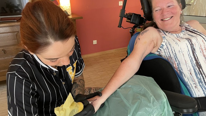 Eine Tätowiererin sticht einer lächelnden Frau ein Tattoo auf den Arm.
