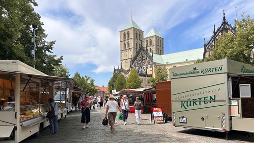 Menschen schlendern über den Wochenmarkt in Münster. Im Hintergrund der St. Paulus-Dom.