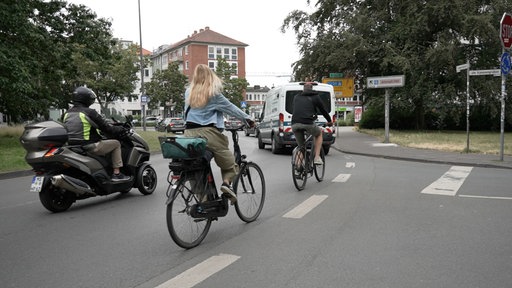 Im Vordergrund fahren zwei Fahrräder und ein Motorradfahrer durch einen Kreisverkehr, vor ihnen fahren Autos.