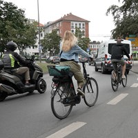 Im Vordergrund fahren zwei Fahrräder und ein Motorradfahrer durch einen Kreisverkehr, vor ihnen fahren Autos.