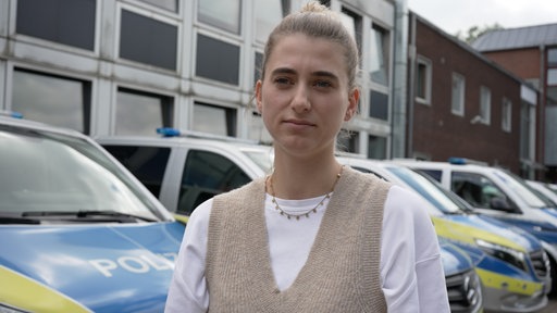 Eine Frau schaut in die Kamera, hinter ihr stehen Polizei-Autos.