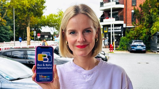 WDR-Reporterin Julia Wulf hält ein Handy mit dem Logo von "Bus und Bahn Münsterland" in der Hand.