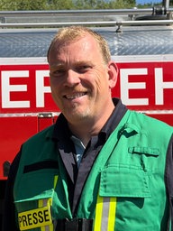 Nico Holtkemper, Leiter der Feuerwehr Ostbevern, steht vor einem Einsatzwagen und lächelt in die Kamera.