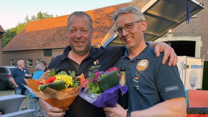 Die beiden Ballonfahrer mit Blumensträußen umarmen sich auf einem Hof