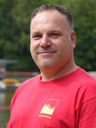 Ein Mann in rotem T-Shirt vor einem Badesee.