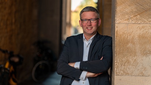 Ein Mann steht mit verschränkten Armen an eine Hauswand gelehnt und lächelt in die Kamera 