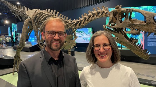Museumsdirektor Jan-Ole Kriegs und Kuratorin Lisa Wong stehen vor einem Saurier-Skelett.