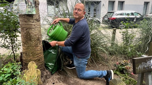ein Mann füllt einen Beutel mit Wasser an einem Baum in einem Blumenbeet