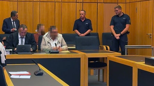 Im Gerichtssaal sitzen zwei Angeklagte. Außerdem zwei polizisten und zwei weitere Personen im Anzug