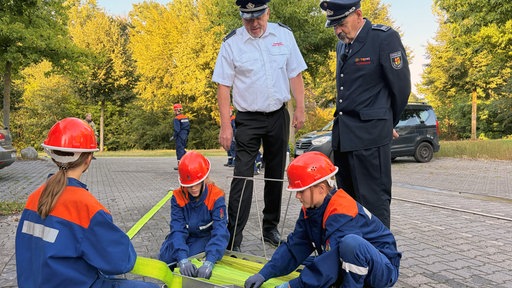 Drei Mädchen in Feuerwehruniform rollen einen Wasserschlauch zusammen, zwei Feuerwehrmänner stehen daneben.