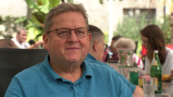 Ein Mann mit Brille und petrolfarbenem Polo-Shirt sitzt an einem Tisch in einer Pflegeeinrichtung. Im Hintergrund sind weitere, sitzende Menschen erkennbar.