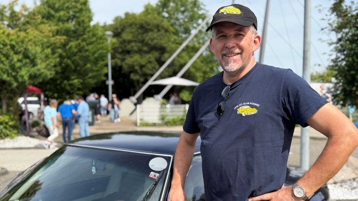 Ein Mann steht mit einer Auto-Kappe und einem dazu passenden Auto-T-Shirt neben einem dunkelblauen Opel GT.