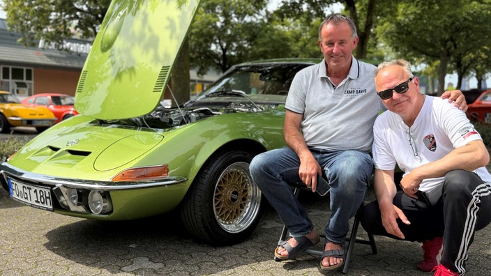 Zwei Männer neben einem grünen Opel GT. Der eine hat seinen Arm über der Schulter des anderen liegen.