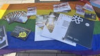 Broschüren und Flyer über LGBTQ und Queerfeindlichkeit liegen auf einem Tisch, der mit einer Regenbogenflagge bedeckt ist.