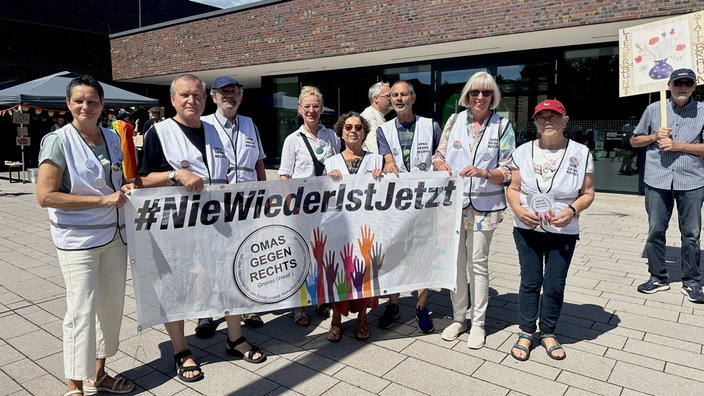 Mehrere Frauen und Männer in weißen Westen stehen in einer Gruppe und halten ein Banner mit der Aufschrift "Nie wieder ist jetzt".