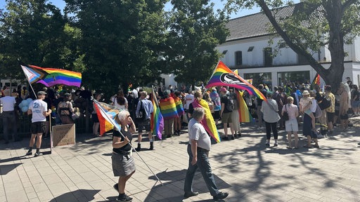Eine große Personengruppe versammelt sichauf einem Platz. Viele von ihnen tragen Regenbogenfahnen.