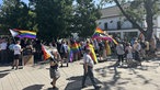 Eine große Personengruppe versammelt sichauf einem Platz. Viele von ihnen tragen Regenbogenfahnen.
