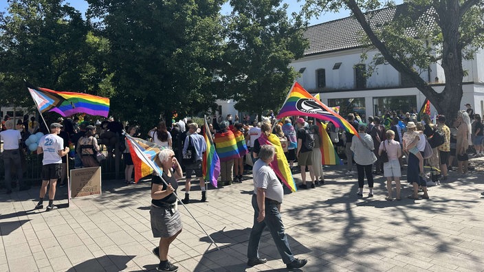 Eine große Personengruppe versammelt sichauf einem Platz. Viele von ihnen tragen Regenbogenfahnen.