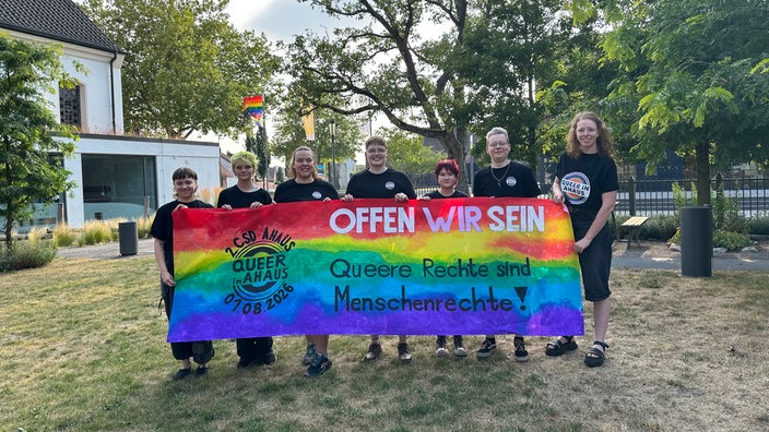 Eine Gruppe aktivistischer Personen, die ein Banner in Regenbogenfarben emporhält: "Offen wir sein. Queere Rechte sind Menschenrechte!"