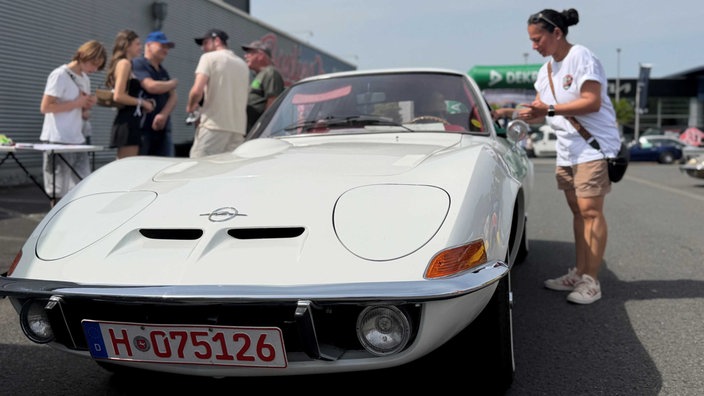 Ein weißer Oldtimer der Marke Opel, daneben spricht eine Frau durch das Autofenster mit dem Fahrer und reicht einen Gegenstand ins Auto.