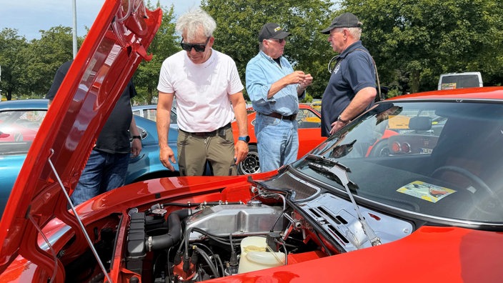 Drei Männer stehen neben einem Auto. Zwei sprechen miteinander, einer schaut unter die Motorhaube des Opel GTs.