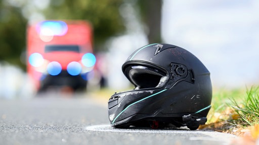 Ein Motorradhelm liegt, mit Kreide umrandet, vor einem Notarztwagen auf einer Landstraße