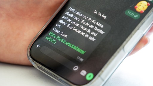 Ein Handy zeigt einen WhatsApp Chat mit folgender betrügerischer Nachricht "Hallo! Könntest du für Klara abstimmen? Sie ist die Tochter meiner engen Freunde, und dieser Sieg beudeutet ihr sehr viel. Vielen Dank." Danach folgt ein Link.