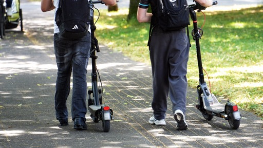 Zwei Menschen schieben jeweils einen E-Scooter neben sich her.